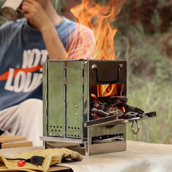 A stainless steel camping stove with a fire burning inside. The stove is sitting on a table with a pair of gloves and a person in the background.